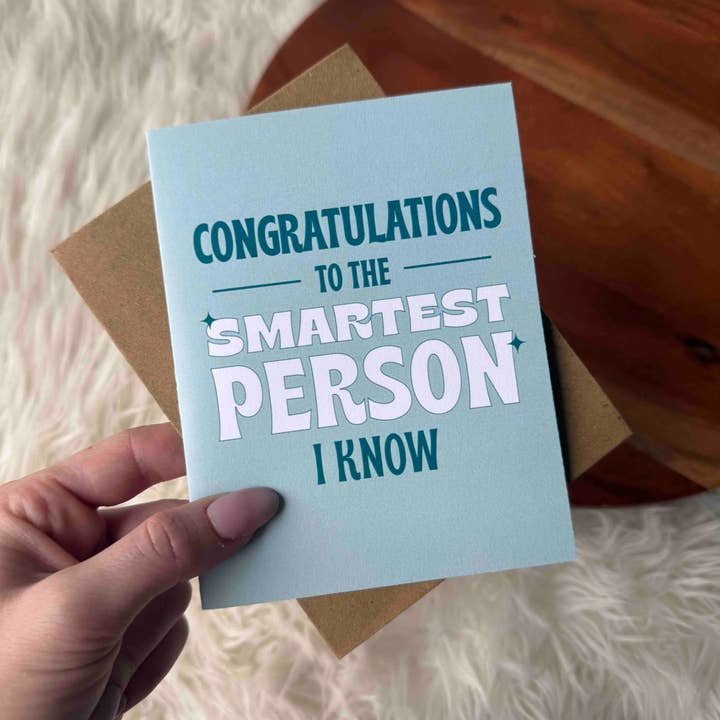 A hand holds the Big Moods "Congrats To the Smartest Person I Know Card" with an envelope on a fuzzy white rug—perfect for celebrating a college graduation.