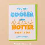 The Cooler/Hotter Birthday Card by And Here We Are features blue and orange text, neon inks, and flames around "hotter" for a bold birthday wish. Hand printed and vibrant—a standout card for any celebration.