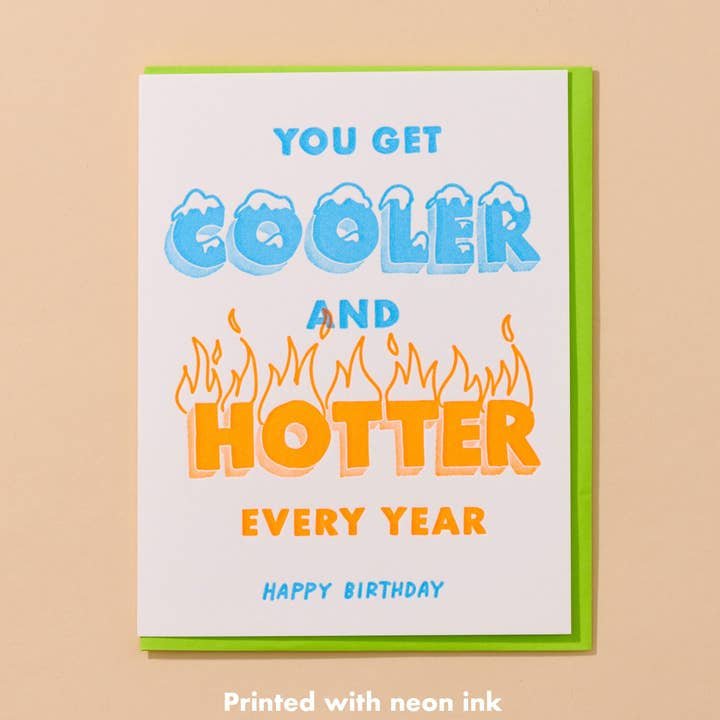 The Cooler/Hotter Birthday Card by And Here We Are features blue and orange text, neon inks, and flames around "hotter" for a bold birthday wish. Hand printed and vibrant—a standout card for any celebration.