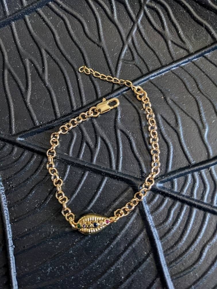 The Coquille D'or Bracelet by Minh Atelier features an 18k gold plated design with an oval shell charm and embedded stones. Nickel free, it adds elegance to any look. Displayed on a textured black surface with geometric patterns.