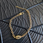 The Coquille D'or Bracelet by Minh Atelier features an 18k gold plated design with an oval shell charm and embedded stones. Nickel free, it adds elegance to any look. Displayed on a textured black surface with geometric patterns.