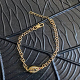 The Coquille D'or Bracelet by Minh Atelier features an 18k gold plated design with an oval shell charm and embedded stones. Nickel free, it adds elegance to any look. Displayed on a textured black surface with geometric patterns.