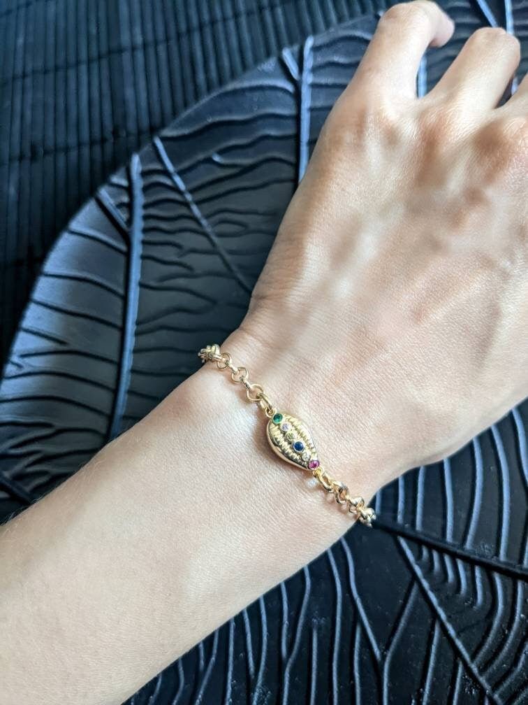 A hand wears the Coquille D'or Bracelet by Minh Atelier—an 18k gold plated piece with a decorative oval charm set with small colored stones—resting on a black textured surface.