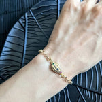 A hand wears the Coquille D'or Bracelet by Minh Atelier—an 18k gold plated piece with a decorative oval charm set with small colored stones—resting on a black textured surface.