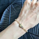 A hand wears the Coquille D'or Bracelet by Minh Atelier—an 18k gold plated piece with a decorative oval charm set with small colored stones—resting on a black textured surface.