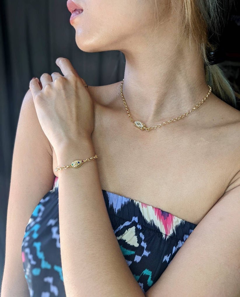 A woman in a patterned strapless dress highlights Minh Atelier’s Coquille D'or Bracelet, an 18k gold plated, nickel-free piece with small diamond-shaped pendants, paired with a matching gold chain necklace.