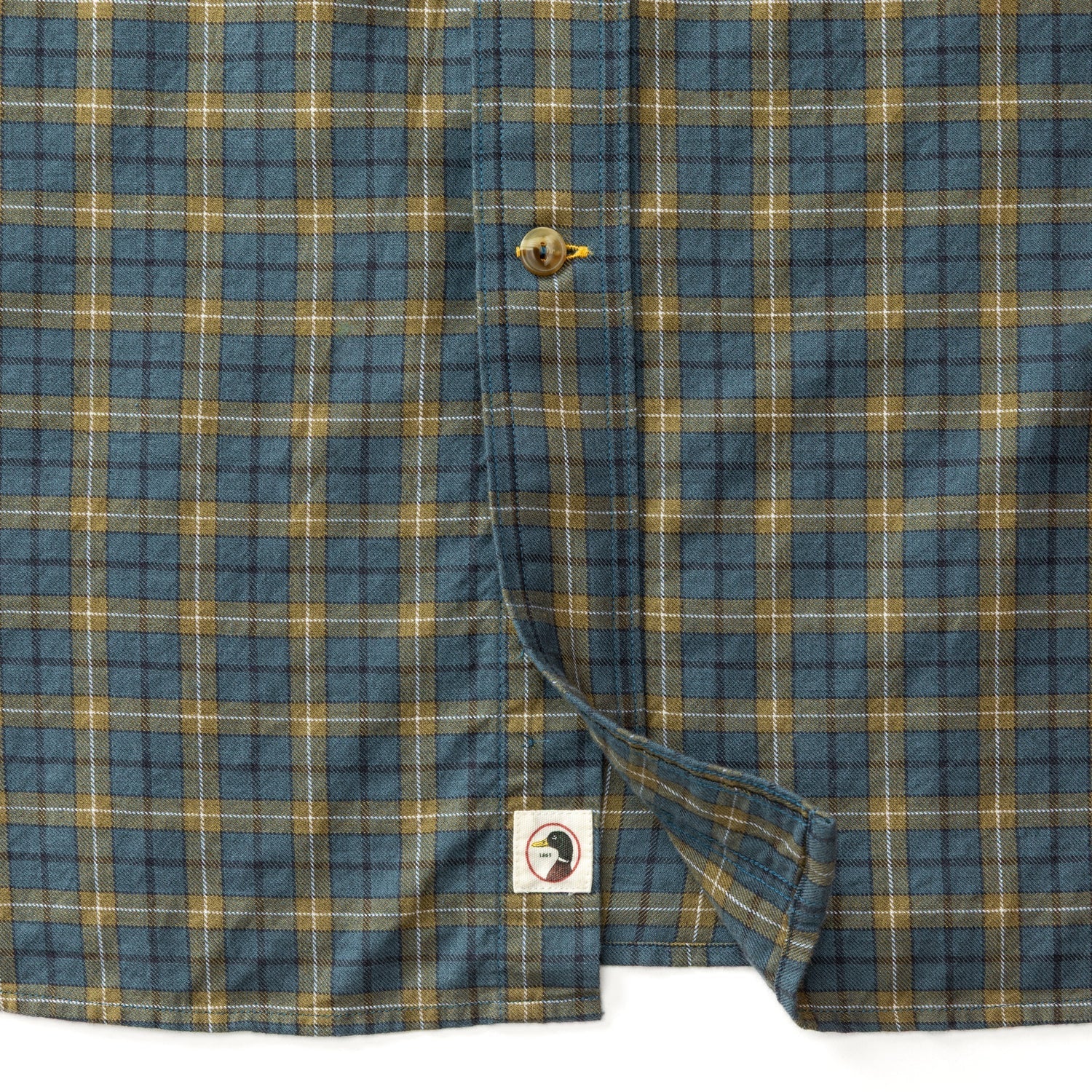 Close-up of the lower part of a Duck Head Cotton Wool Sport Shirt in Orion Blue, yellow, and brown Mercer Plaid, featuring a button and a small bird logo tag near the hem.