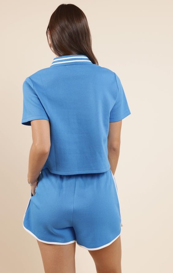 A person with long brown hair is seen from the back, wearing a small Courtside Striped Collar Polo and shorts by Sadie And Sage, featuring blue fabric with white trim in a comfy cotton-polyester blend for women.