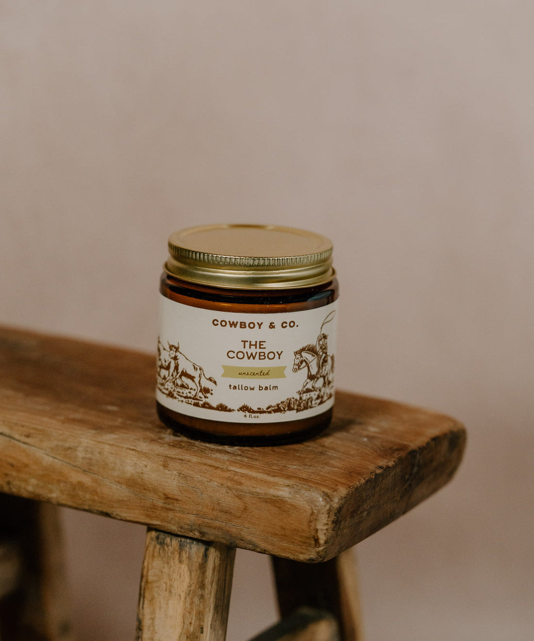 A glass jar of Cowboy & Co. COWBOY Unscented Tallow Moisturizer with a gold lid sits on a rustic wooden stool against a neutral background.