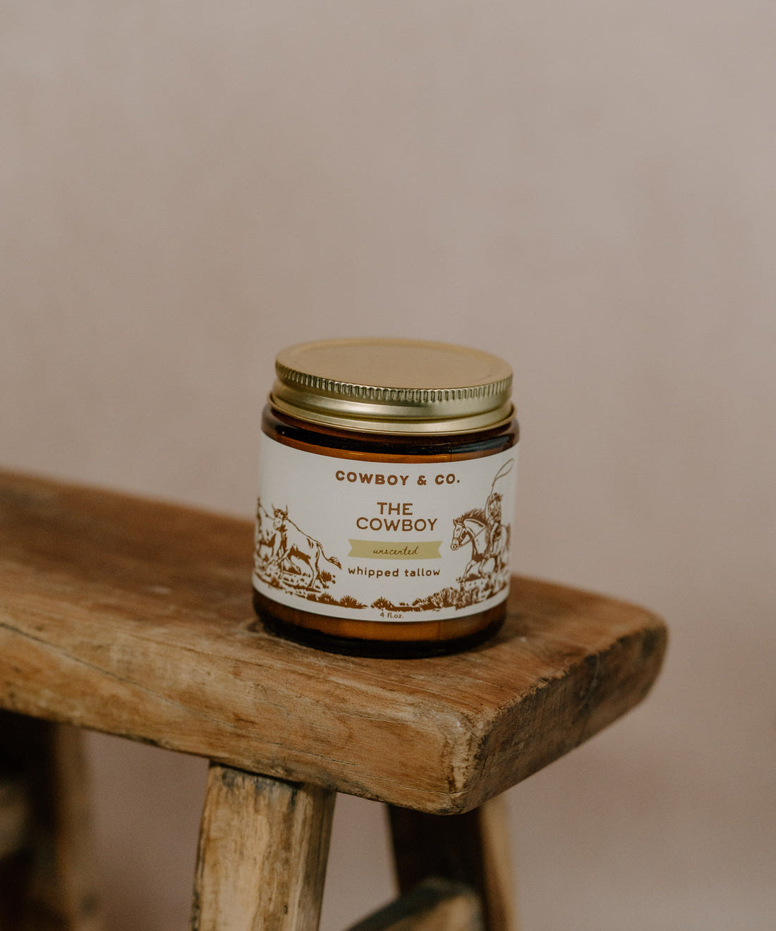 A jar of COWBOY | Unscented Tallow Moisturizer by Cowboy & Co. rests on a rustic wooden stool, its label visible against a neutral background.