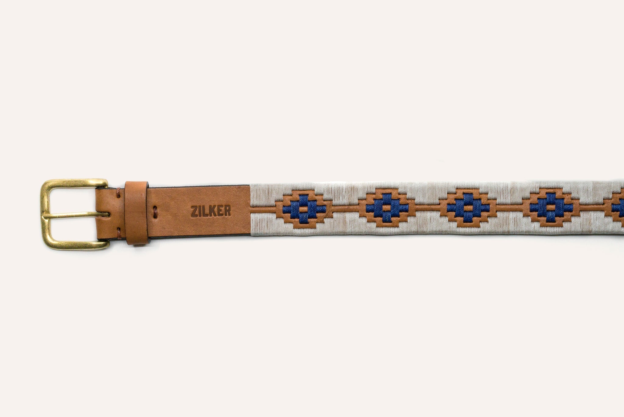 The Cowboy by Zilker Belts is a hand-stitched belt made from premium Argentine leather, with a brass buckle, blue and brown geometric pattern, and "ZILKER" stamped near the buckle.