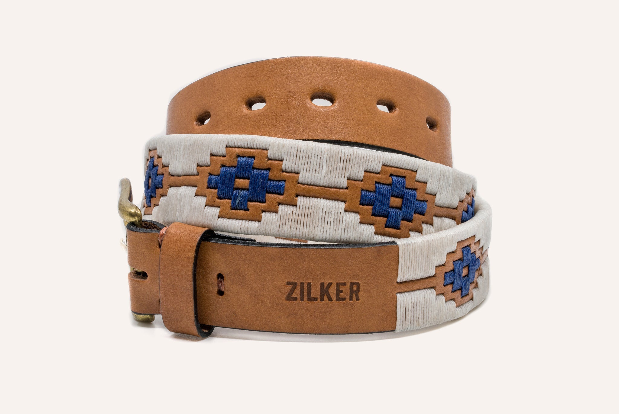 The Cowboy by Zilker Belts features brown Argentine leather with "ZILKER" stamped near the buckle and a white and blue hand-stitched geometric pattern. The belt is displayed coiled in a loop.