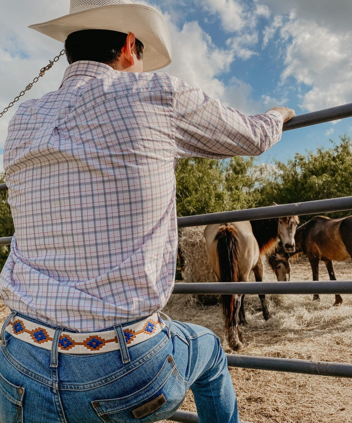 Wearing a cowboy hat, plaid shirt, and the Cowboy by Zilker Belts, someone leans on a metal fence watching horses in a corral under partly cloudy skies.