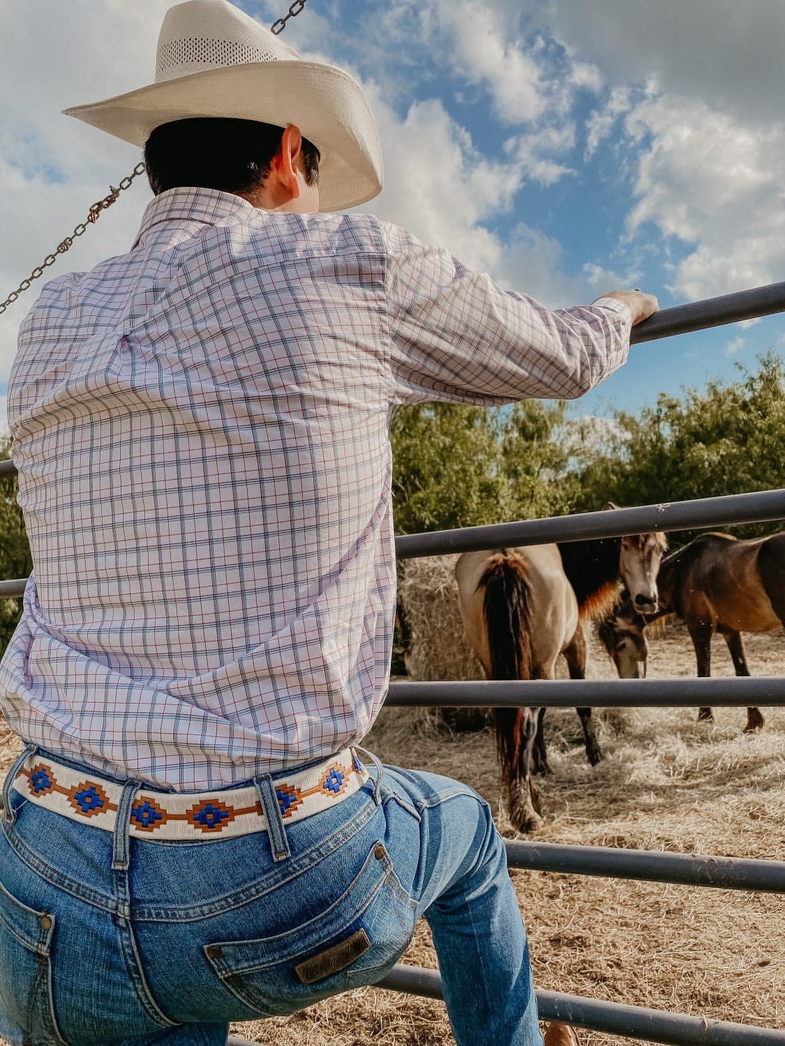 Wearing a cowboy hat, plaid shirt, and the Cowboy by Zilker Belts, someone leans on a metal fence watching horses in a corral under partly cloudy skies.