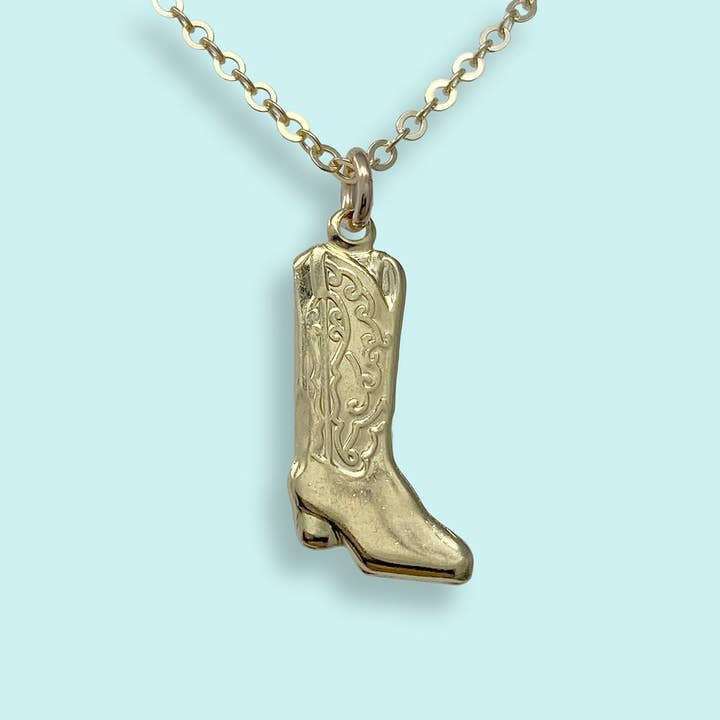 The Cowgirl Boot Necklace by Ornamental Things features a gold cowboy boot pendant with engraved designs, showcased against a light blue background.