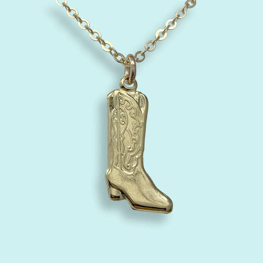 The Cowgirl Boot Necklace by Ornamental Things features a gold cowboy boot pendant with engraved designs, showcased against a light blue background.
