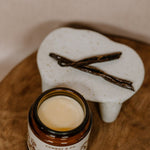 An open jar of Cowboy & Co. COWGIRL Vanilla Infused Tallow balm for sensitive skin sits beside a ceramic dish with two vanilla beans on a wooden surface.