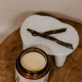 An open jar of Cowboy & Co. COWGIRL Vanilla Infused Tallow balm for sensitive skin sits beside a ceramic dish with two vanilla beans on a wooden surface.