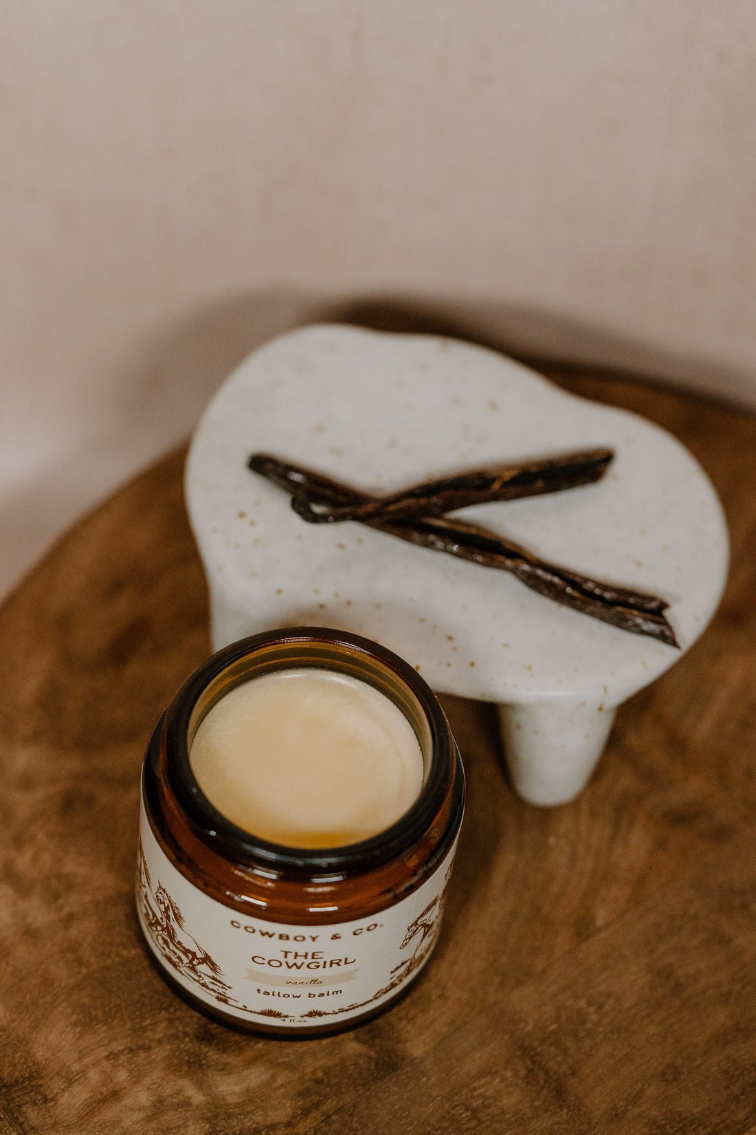 An open jar of Cowboy & Co. COWGIRL Vanilla Infused Tallow balm for sensitive skin sits beside a ceramic dish with two vanilla beans on a wooden surface.