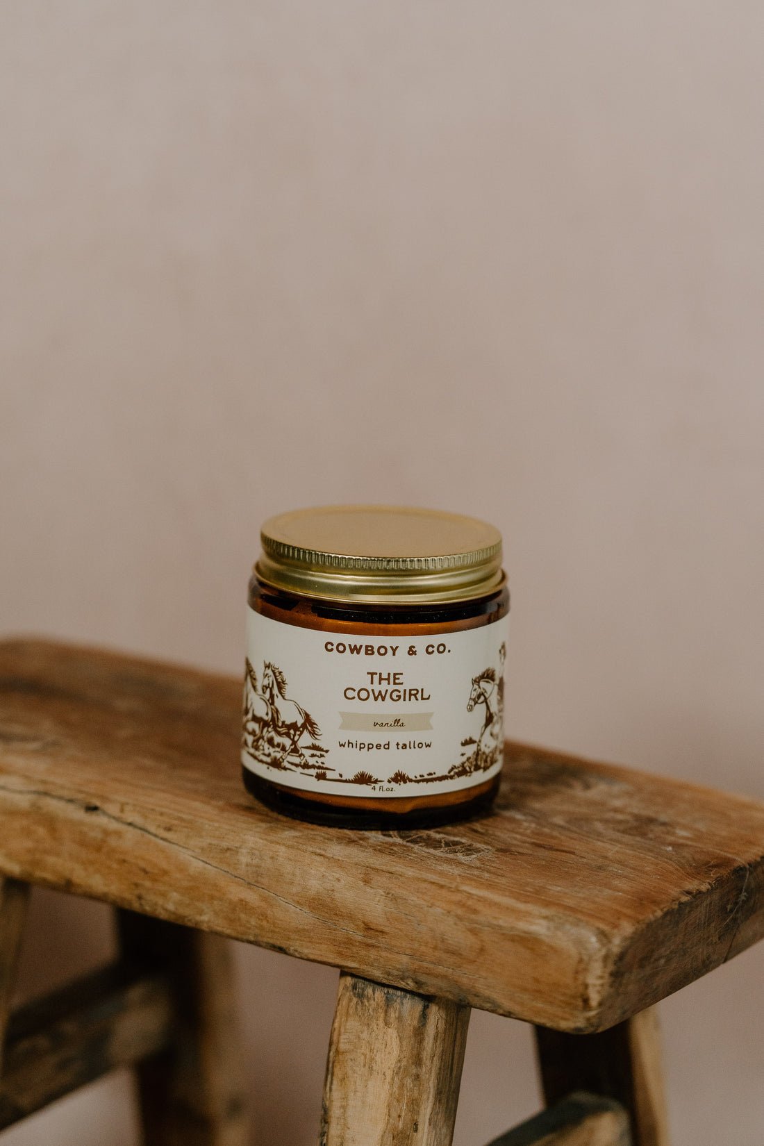 A glass jar labeled "COWGIRL | Vanilla Infused Tallow" by Cowboy & Co. rests on a rustic wooden surface against a plain background—ideal for sensitive skin and made with grass-fed beef tallow.
