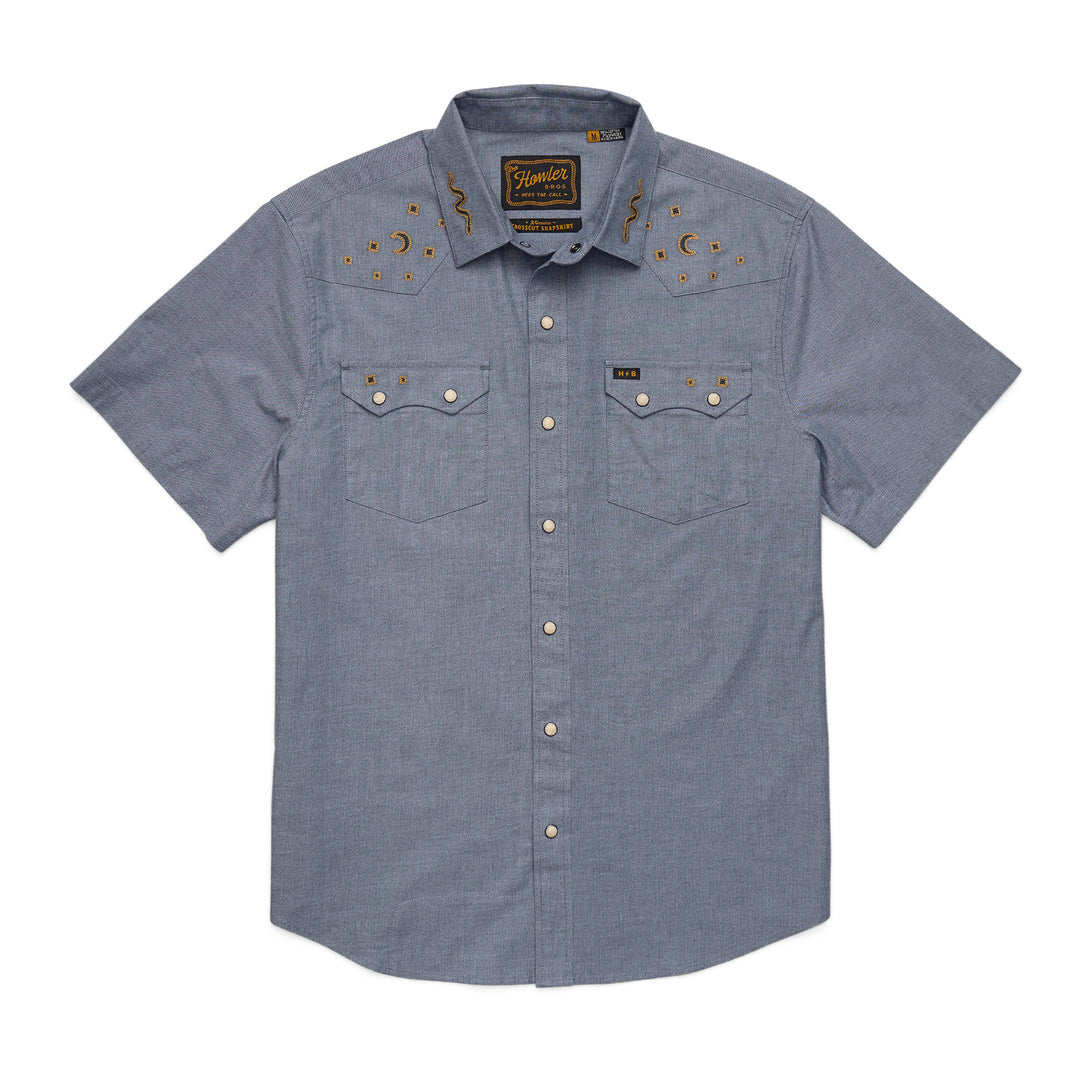 The Howler Brothers Crosscut Deluxe Snapshirt is a short-sleeve, button-up blue chambray with pearl snaps, two chest pockets, custom embroidery, and Western-style shoulder detailing.