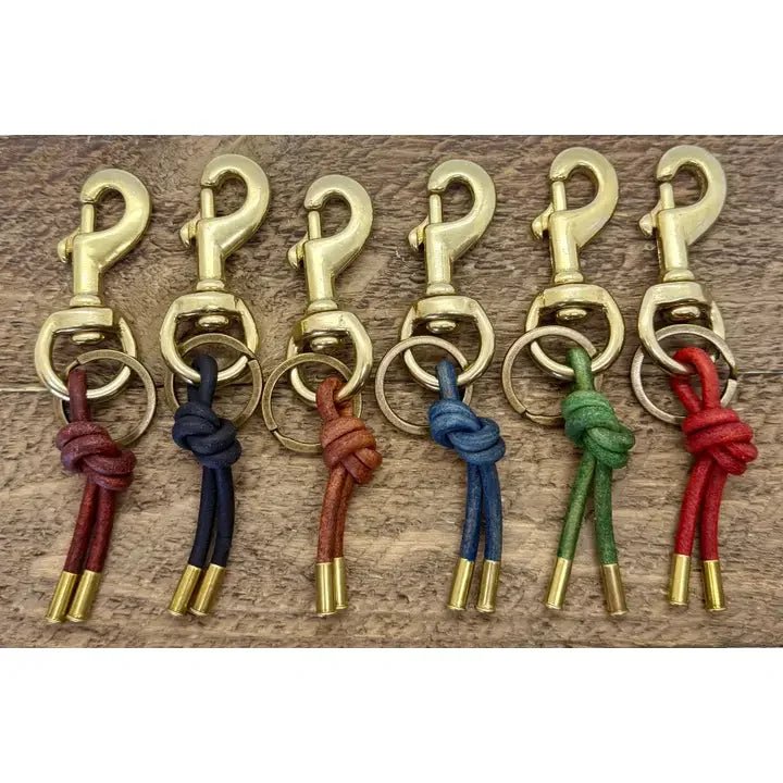 Six Cuatro Keychain hooks by Tres Cuervos, each with a leather cord in black, brown, red, blue, green, or tan, are lined up on a wooden surface.