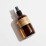 A brown glass bottle labeled “No.11 Amber & Moss Room & Linen Spray” by P.F. Candle Co., phthalate-free, with a black spray nozzle and its shadow on a white surface.