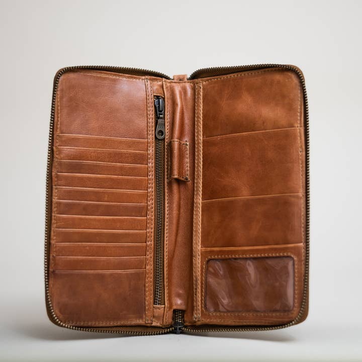The Stash Passport Wallet in caramel features multiple card slots, a zippered compartment, and a clear ID window, shown open against a plain light background.