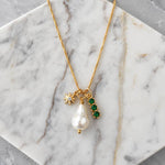Mesa Blue’s Pearl, Cz Emerald, Star Charm Necklace features 18K gold plating and three pendants—a freshwater pearl, a pavé CZ star charm, and a trio of green gemstone accents—elegantly presented on a white marble surface.