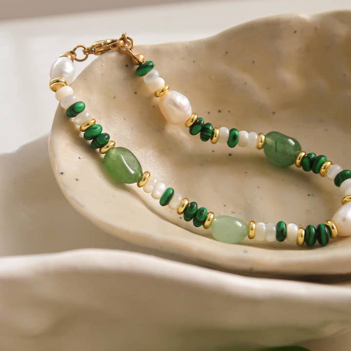 A Faye Bohemian Stone and Bead Bracelet by Flanerie by MAIQ featuring green stone beads, natural pearls, and 18K gold plated spacers, resting in a curved ceramic dish.