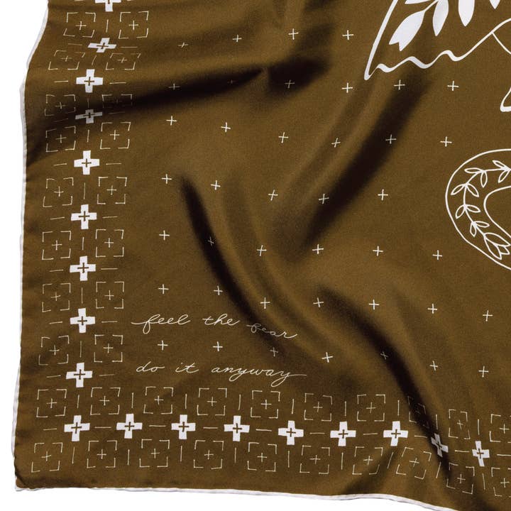 The Silk Scarf - ‘Feel the Fear’ from Jenni Earle features a brown fabric adorned with white geometric patterns, a wing motif, and the inspiring phrase “feel the fear do it anyway.” Drawing on western design influences, this square neck and hair scarf makes a bold and meaningful statement accessory.
