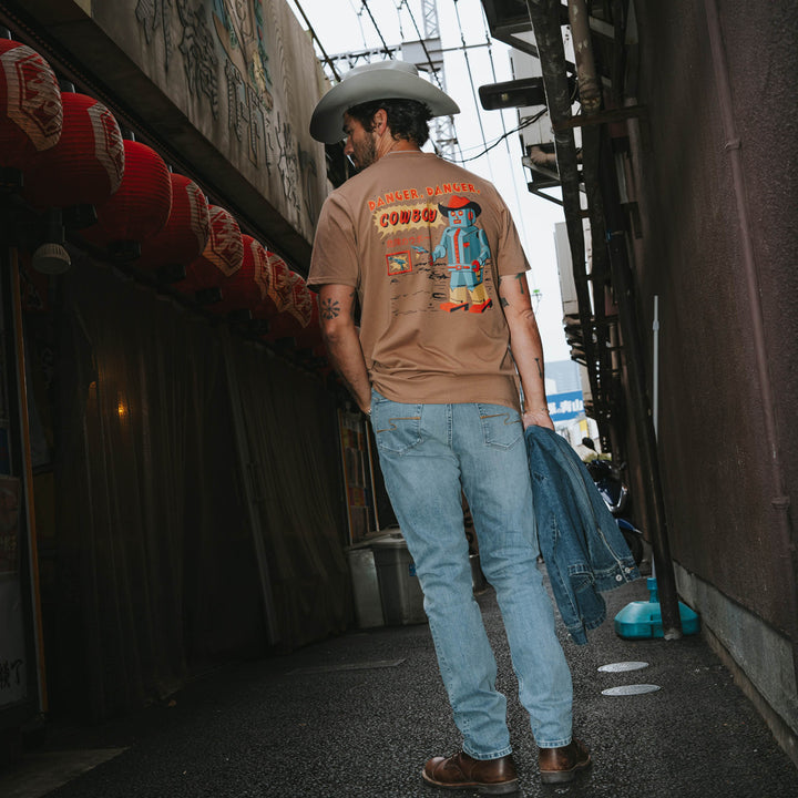 A man in jeans, a cowboy hat, and the Danger Cowboy T-Shirt by Sendero Provisions Co. stands in a narrow alley holding a denim jacket, with red lanterns hanging to his left.
