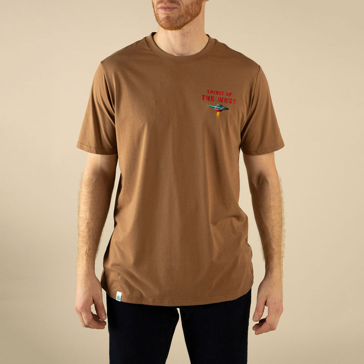 A person wears the Sendero Provisions Co. Danger Cowboy T-Shirt, a soft cotton brown tee with "Spirit of the West" and a small chest graphic, standing against a beige background.