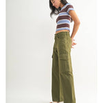Woman in profile wears a striped crop top, JBD Dark Olive Cargo high-rise pants, and white heeled sandals against a plain white background.