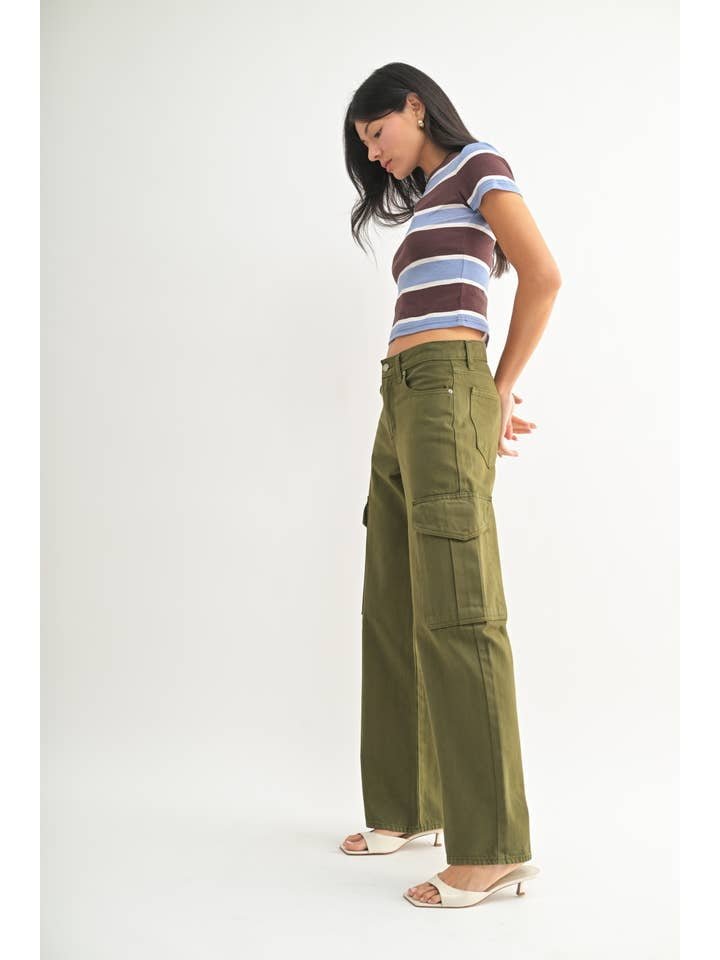 A woman stands sideways in a striped short-sleeve shirt, white heeled sandals, and JBD’s Dark Olive Cargo jeans against a plain white background.