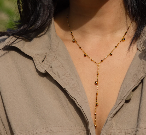 A person in a beige collared shirt wears the Dax Lariat Necklace by Peter and June, a delicate 18k gold plated piece with small round beads, photographed from the shoulders up.