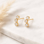 A pair of Mesa Blue Cz Star Climber Stud Earrings, featuring 18K gold plating and three clear gemstones each, displayed on a white marble surface next to a pampas grass stem.