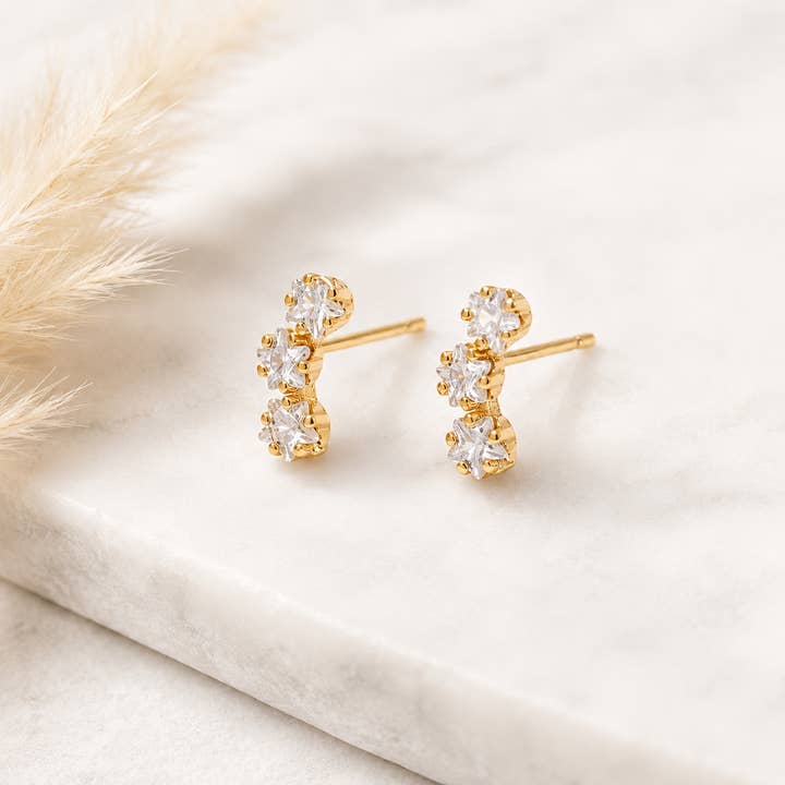 A pair of Mesa Blue Cz Star Climber Stud Earrings, featuring 18K gold plating and three clear gemstones each, displayed on a white marble surface next to a pampas grass stem.