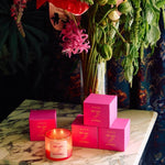 A lit De Las Flores | Roen coconut soy wax candle and four pink Roen boxes are arranged on marble before a vase of assorted flowers, inspired by the vibrant Mexico City flower market.