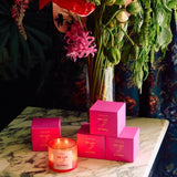 A lit De Las Flores | Roen coconut soy wax candle and four pink Roen boxes are arranged on marble before a vase of assorted flowers, inspired by the vibrant Mexico City flower market.