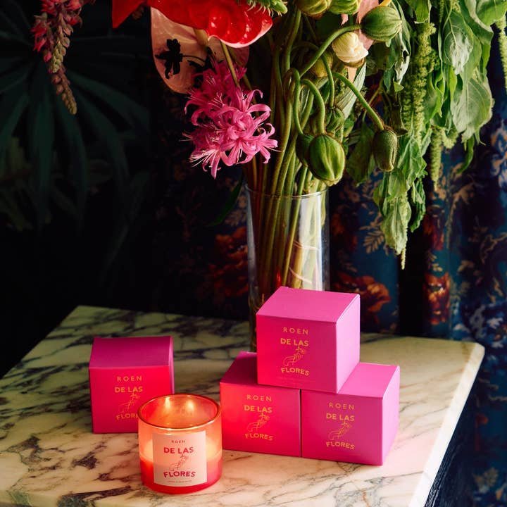 A lit De Las Flores | Roen coconut soy wax candle and four pink Roen boxes are arranged on marble before a vase of assorted flowers, inspired by the vibrant Mexico City flower market.