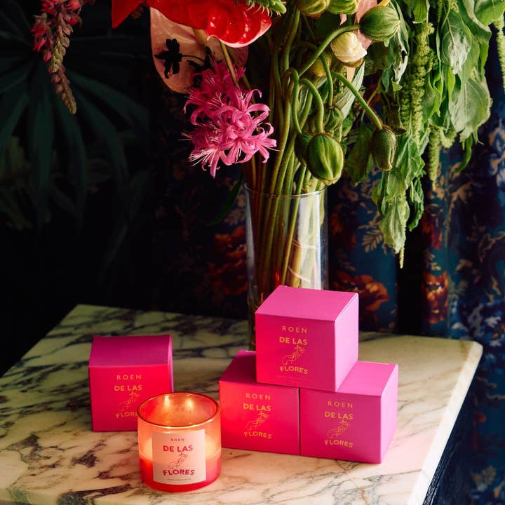 A lit Roen De Las Flores coconut soy wax candle and five pink boxes labeled “De Las Flores | Roen” are displayed on marble before a vase of assorted flowers, capturing the vibrant spirit of a Mexico City flower market.
