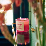 A pink glass "De Las Flores | Roen" candle by Roen, crafted from coconut soy wax with two lit wicks, glows on a reflective surface near a window amid blurred flowers—evoking the lively Mexico City flower market.