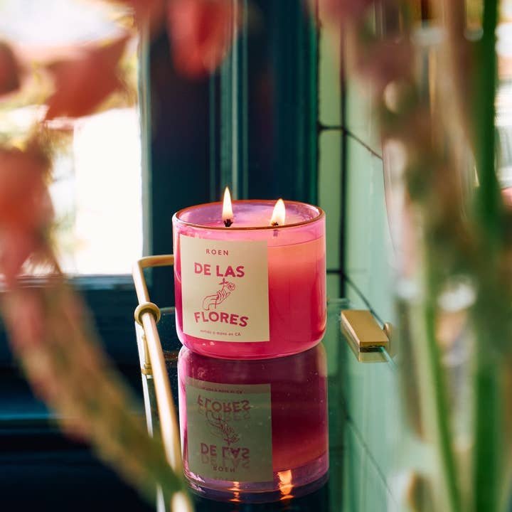 A pink glass "De Las Flores | Roen" candle by Roen, crafted from coconut soy wax with two lit wicks, glows on a reflective surface near a window amid blurred flowers—evoking the lively Mexico City flower market.
