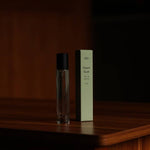 A clear glass bottle labeled "Desert Kush Eau De Parfum | dilo" is placed next to a light green dilo box, both resting on a wooden surface against a dark background.