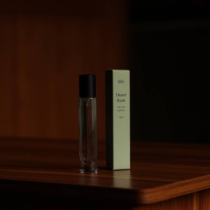 A small glass bottle of Desert Kush Eau De Parfum by dilo sits beside its light green box on a wooden surface, the fragrance highlighted against a dark background.