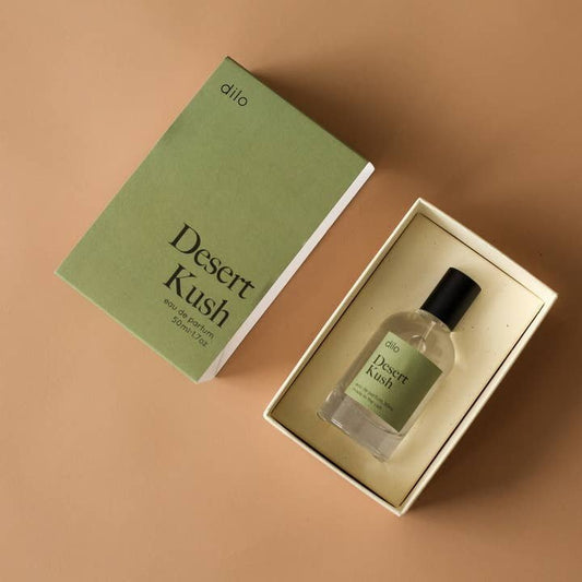 A box labeled "Desert Kush Eau De Parfum | dilo" sits next to an open box containing a matching bottle of Desert Kush Eau De Parfum by dilo on a beige surface.