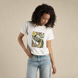 A person in the Sendero Provisions Co. Desert Mark Boxy Tee—a white shirt featuring a colorful cactus and flower graphic—stands against a plain beige background, looking down with one hand in their jeans pocket.