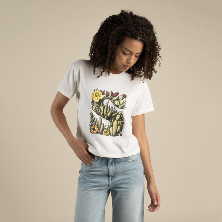 A person in the Sendero Provisions Co. Desert Mark Boxy Tee—a white shirt featuring a colorful cactus and flower graphic—stands against a plain beige background, looking down with one hand in their jeans pocket.