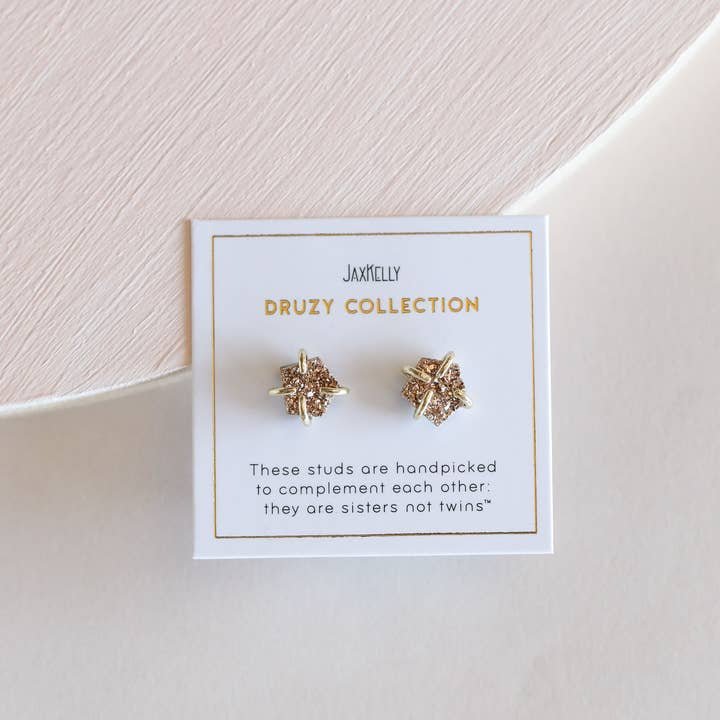 JaxKelly’s Druzy Prong Rose Gold Earrings feature shimmering gold druzy stones, displayed on a white card with “sisters not twins.” Hypoallergenic for comfortable, everyday wear. From the JaxKelly Druzy Collection.